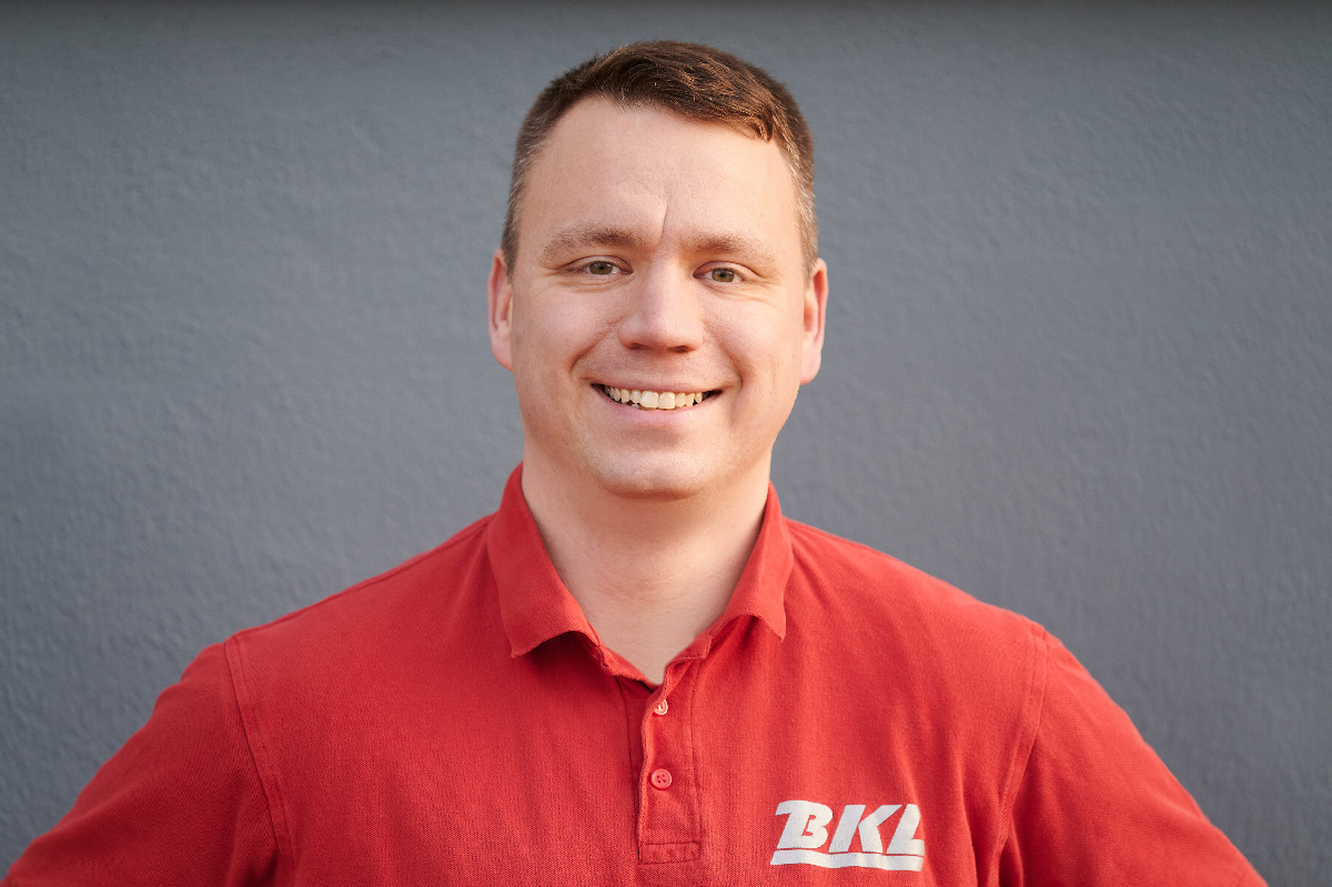 Ein Mann mit einem roten BKL-Poloshirt steht vor einer schlichten grauen Wand und lächelt in die Kamera.
