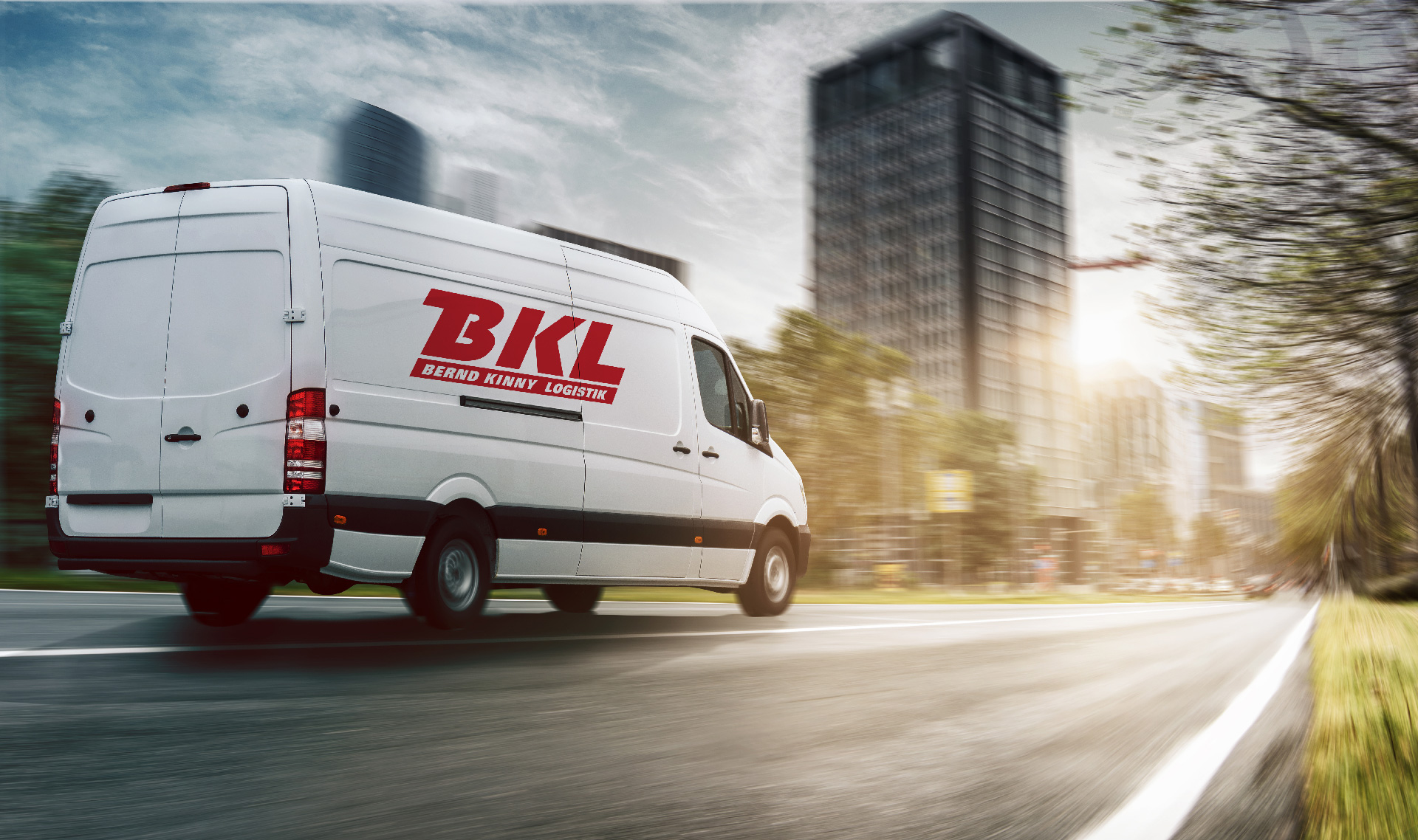 Ein weißer Lieferwagen mit dem Schriftzug "BKL Bernd Klink Logistik" fährt eine Stadtstraße entlang, im Hintergrund sind hohe Gebäude und Bäume zu sehen.