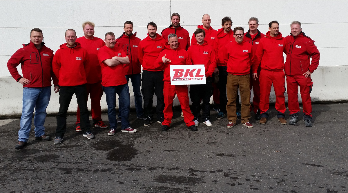 Eine Gruppe von sechzehn Männern in roten Uniformen steht im Freien auf dem Bürgersteig, eine Person in der Mitte hält ein Schild mit der Aufschrift "BKL.