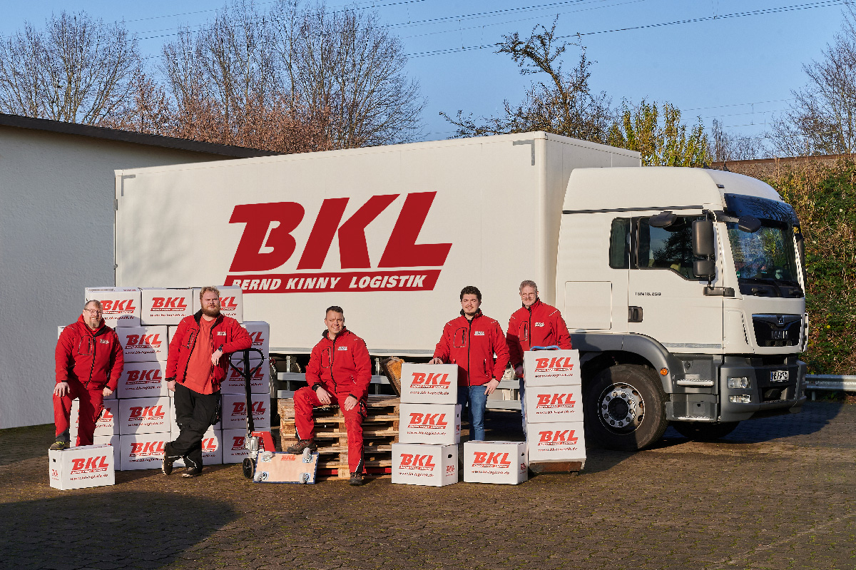 Fünf Personen in roten Jacken posieren mit Kartons der Marke BKL und einem großen BKL-Lieferwagen, der draußen auf einer gepflasterten Fläche steht.